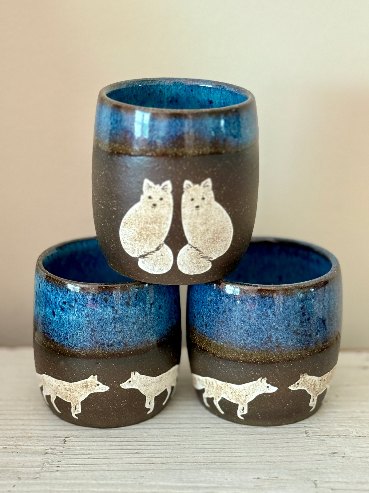 A hand holding a handmade ceramic cup with a dark blue glaze, featuring two white owl silhouettes and a white crescent moon decorated with black speckles, set against a blurred green background.