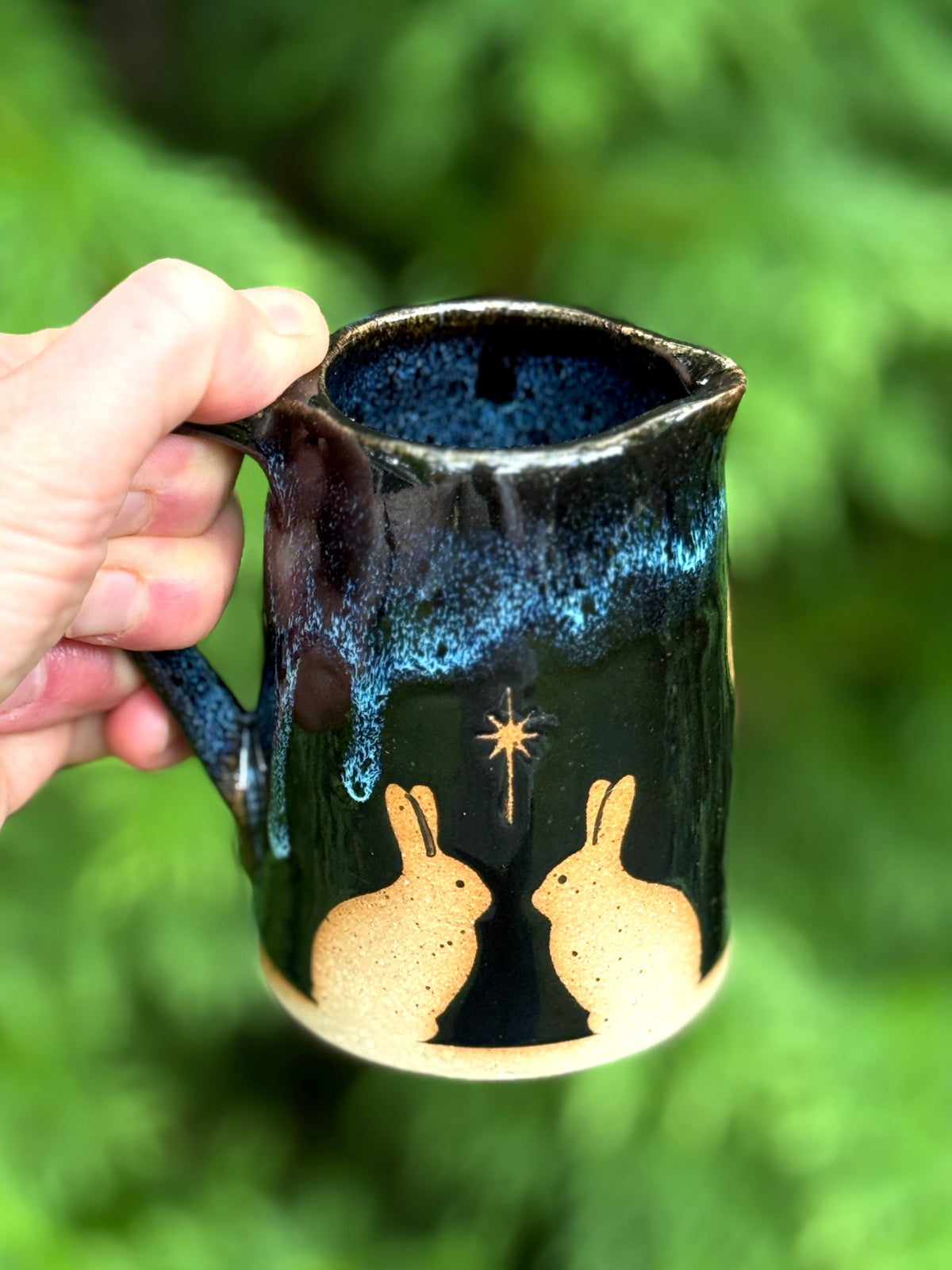 A hand holds a handmade ceramic creamer mug with a dark blue and black glaze, featuring gold rabbit silhouettes and a star design, set against a blurred green background.