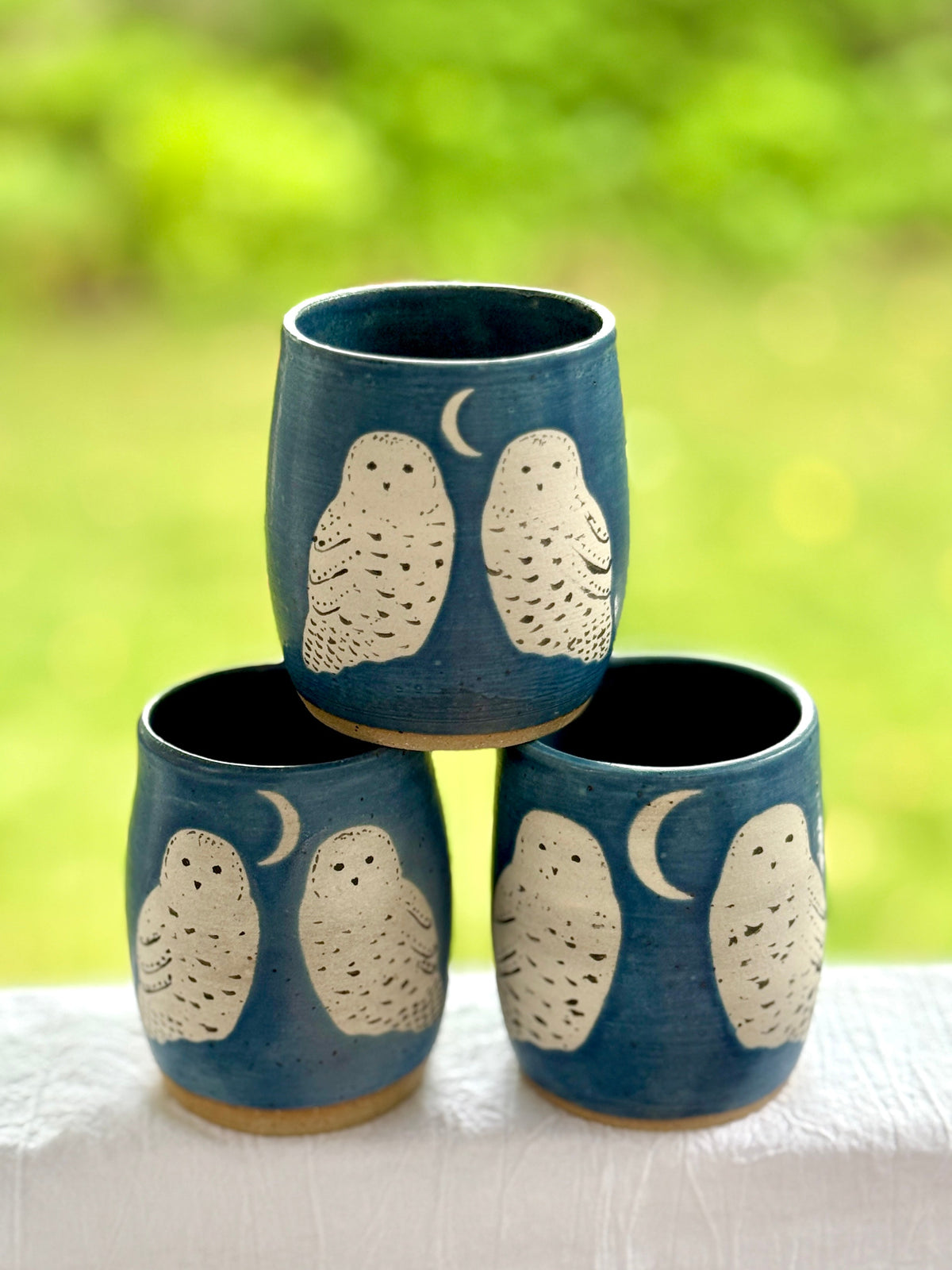 Three ceramic cups stacked in a pyramid, decorated with cream-colored owl and crescent moon motifs on a dark blue glaze, set against a blurred green outdoor background.