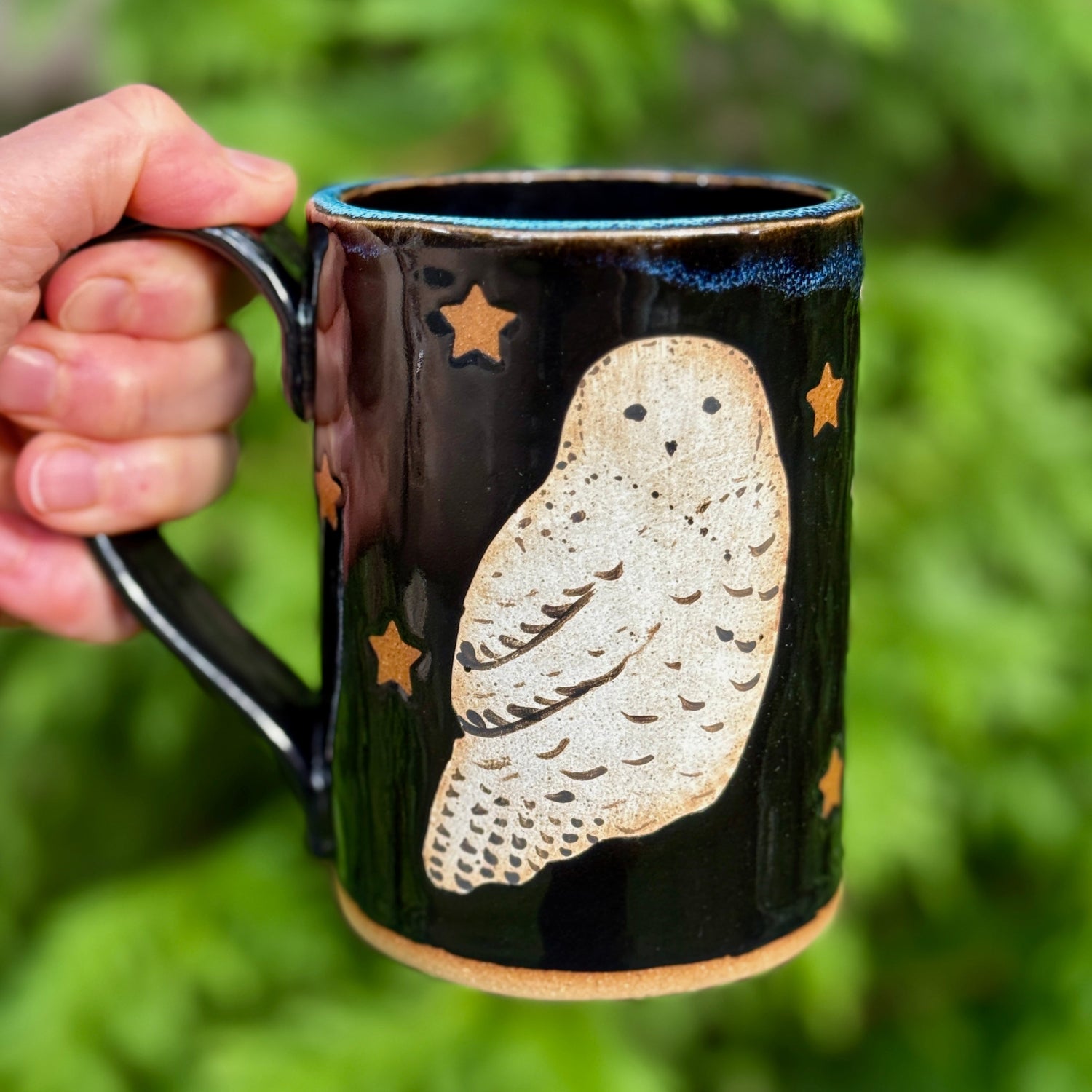 A hand holds a black ceramic mug featuring a hand-painted beige owl and gold stars, set against a blurred green fern background.