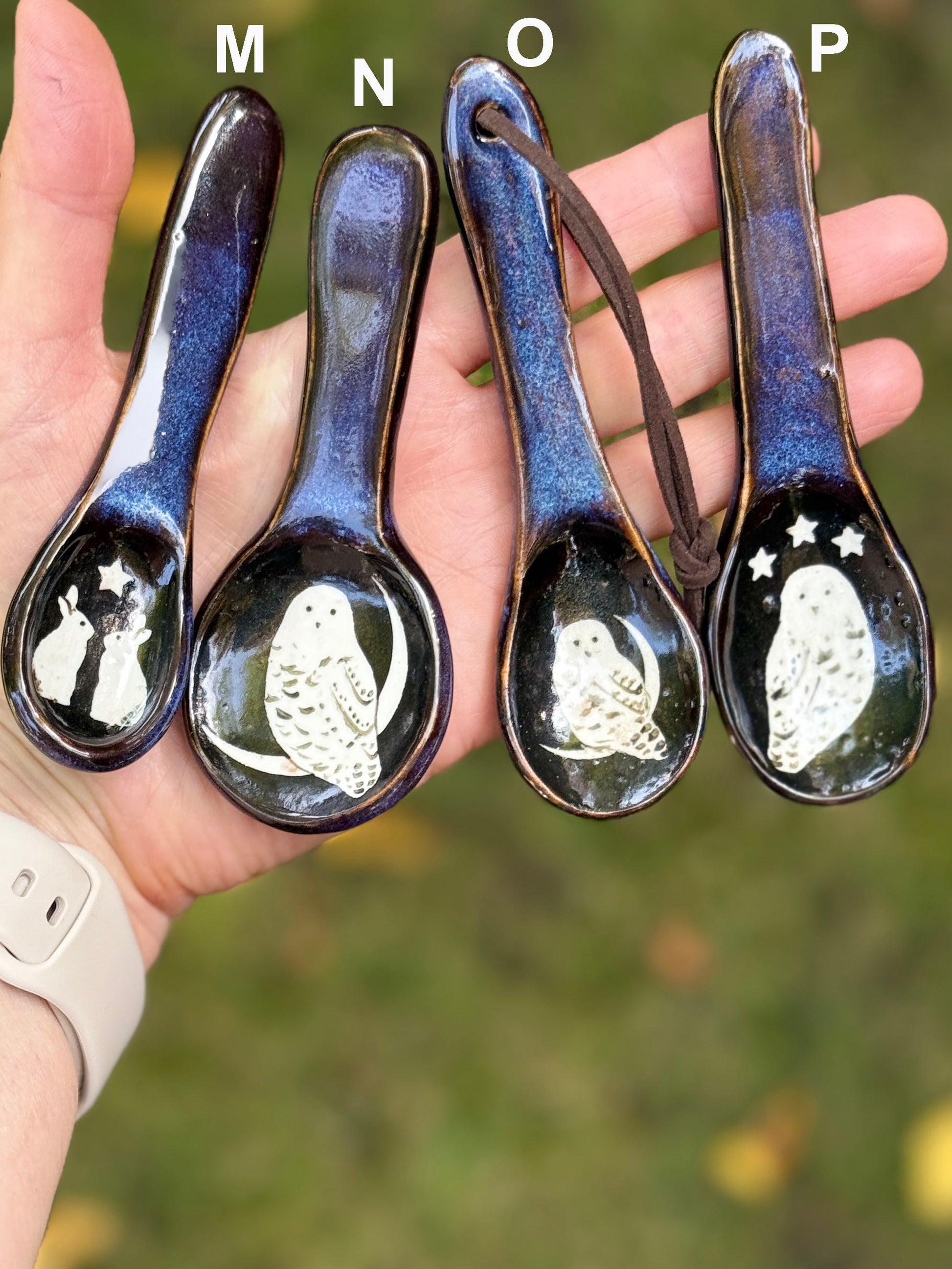 Four ceramic spoons with illustrated designs held in a hand against a blurred natural background.