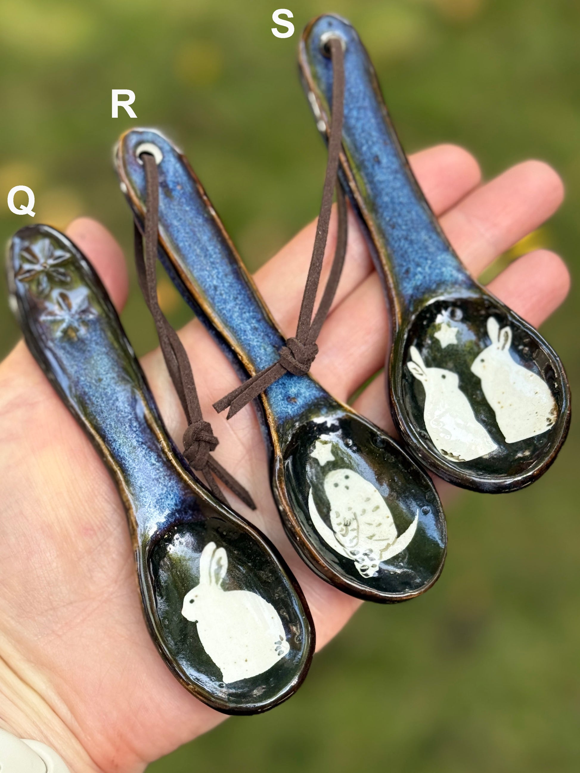 Three ceramic spoons with owl and rabbit designs on a hand against a blurred natural background